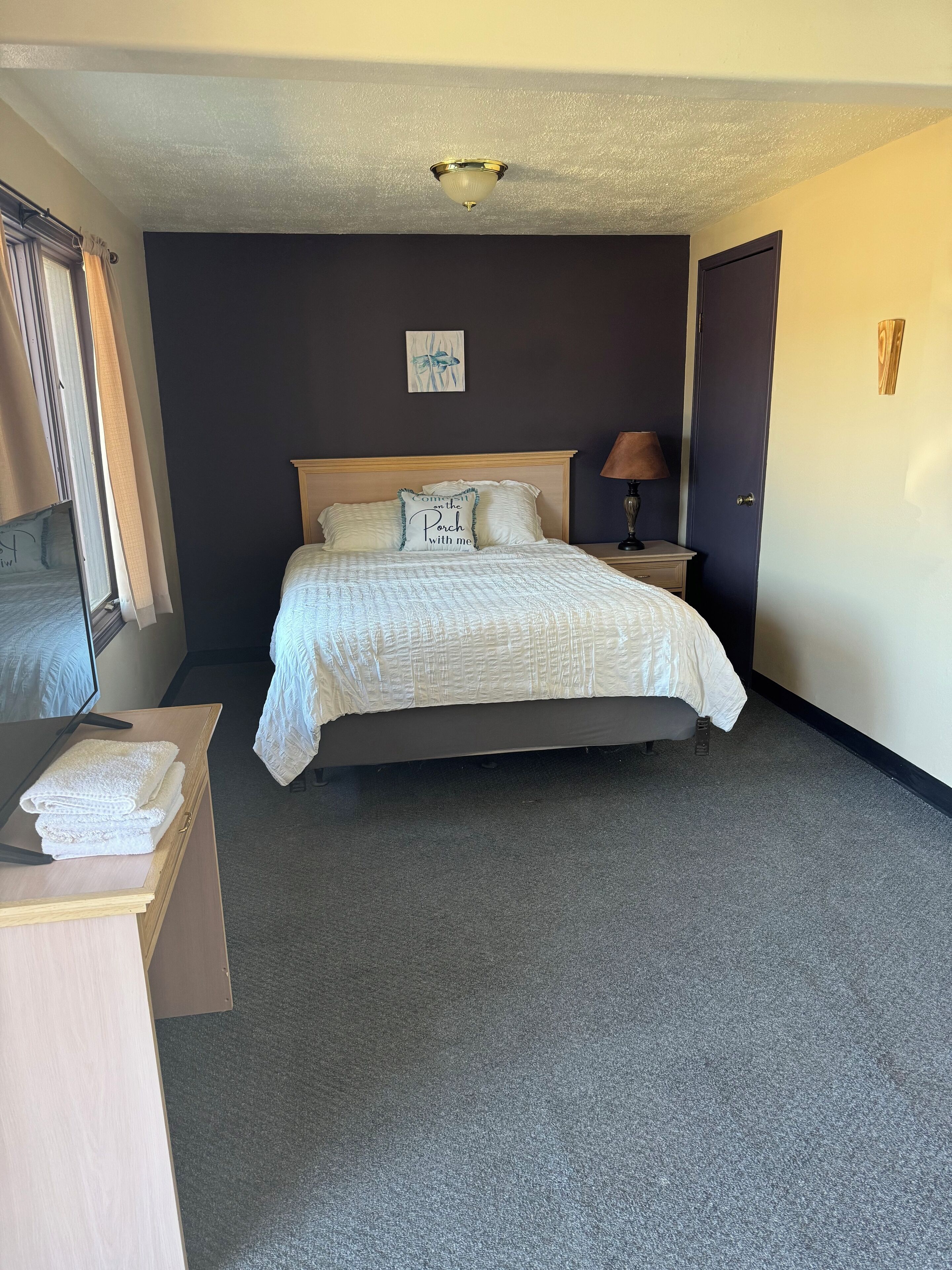 A neatly made bed with a white comforter and pillows, a bedside table with a lamp, a small artwork on the wall, and a door to the right.