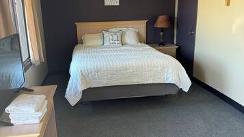 A neatly made bed with a white comforter and pillows, a bedside table with a lamp, a small artwork on the wall, and a door to the right.