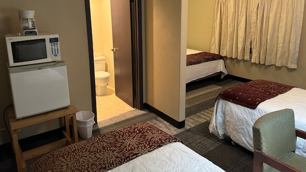 A hotel room with a microwave on a small table, a bed with a patterned bedspread, a second bed with a plain bedspread, a bathroom with a toilet, and a shower curtain.