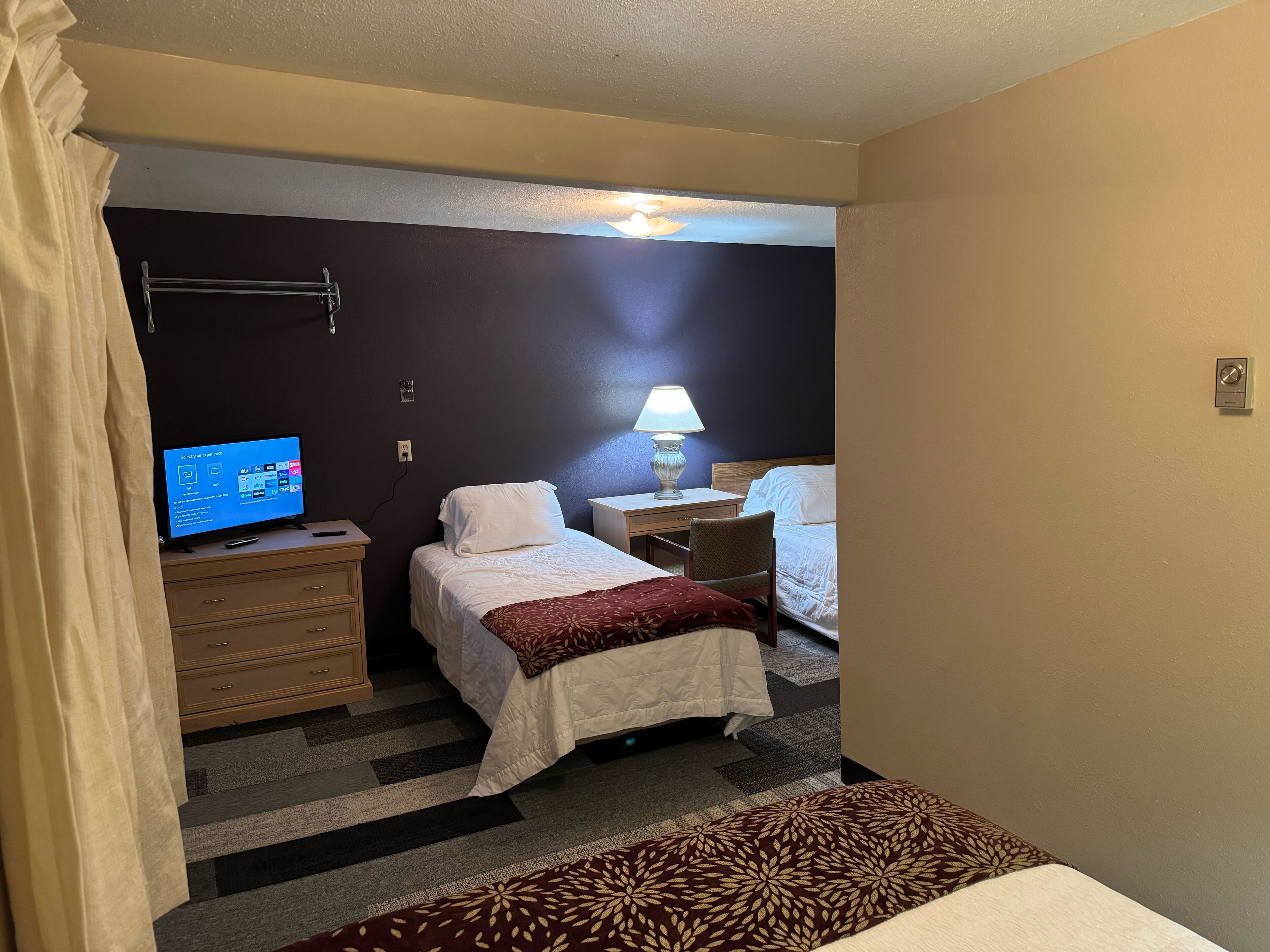 A hotel room with two beds, a desk, a chair, a TV, and a lamp.