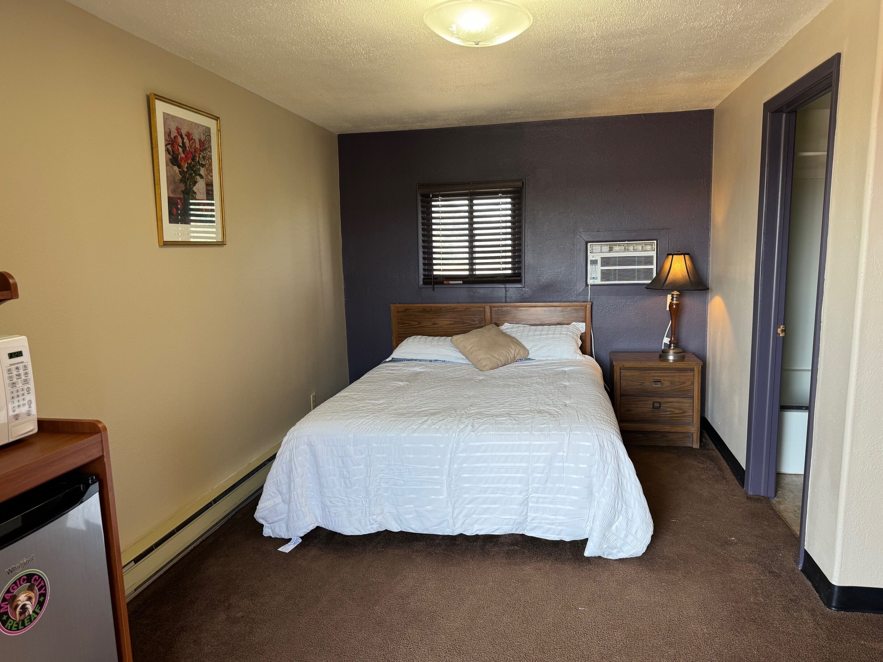 A bedroom with a bed, bedside tables, a wall-mounted air conditioner, a window with blinds, and a door.