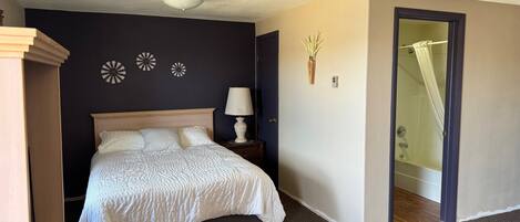 A bedroom with a bed, bedside table, lamp, wall decor, and a shower area.