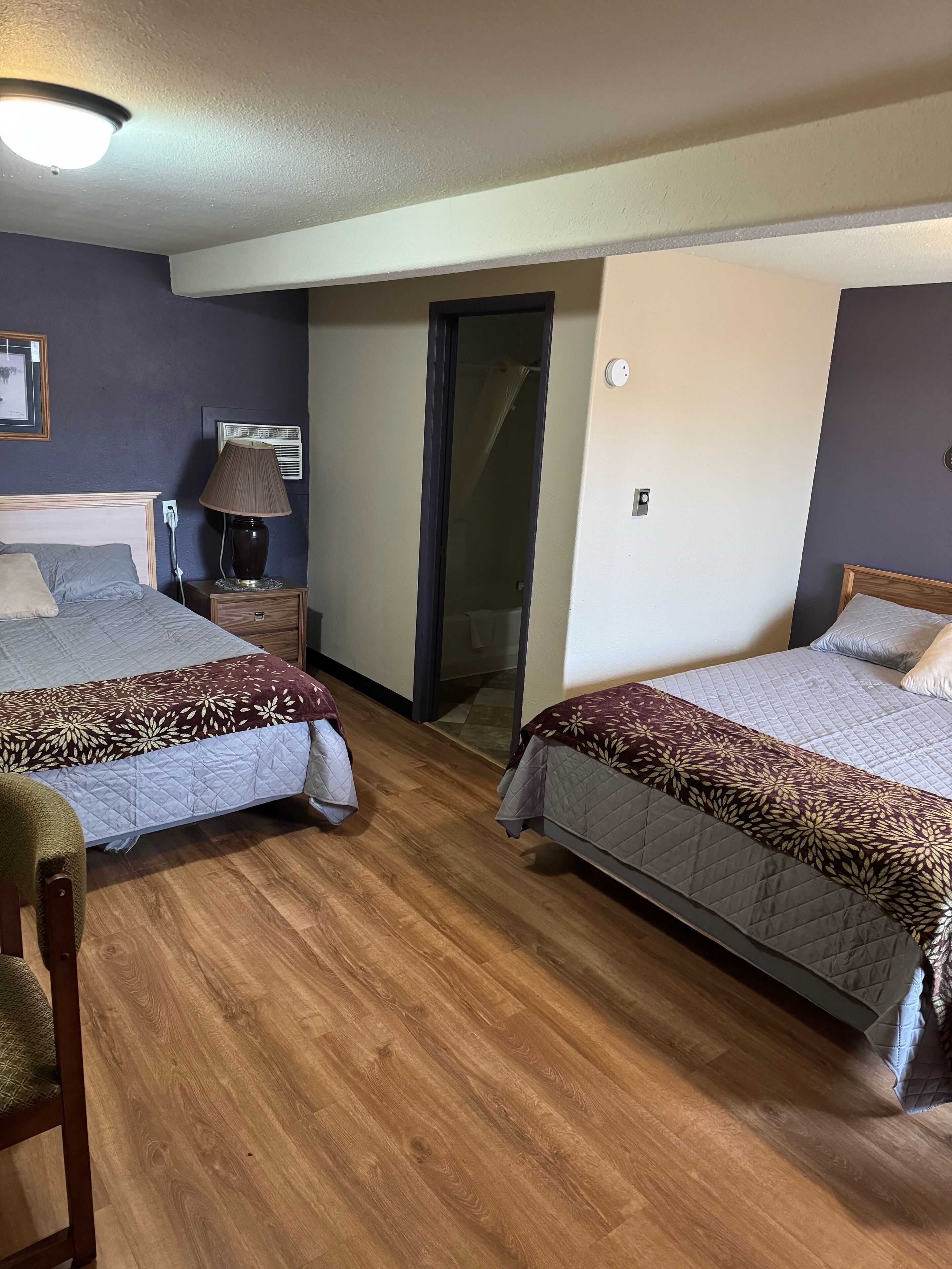 A hotel room with two beds, a nightstand with a lamp, a chair, and a door leading to a bathroom.