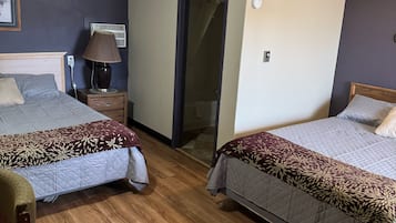 Habitación de hotel con dos camas, una mesita de noche con lámpara, una silla y una puerta que da a un baño.