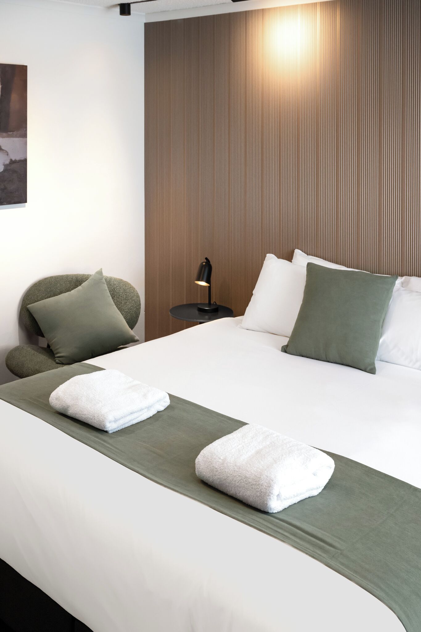 A hotel room with a neatly made bed, a green throw blanket, and a white towel.