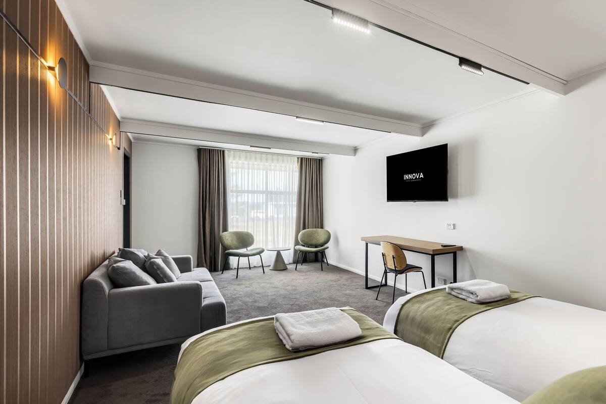 A modern hotel room with a sofa, two beds, a desk, and a TV.