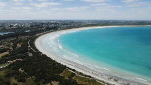 An aerial view of a coastal area with a sandy beach, a clear blue sea, and a distant cityscape. - Innova Motel Warrnambool (Warrnambool)