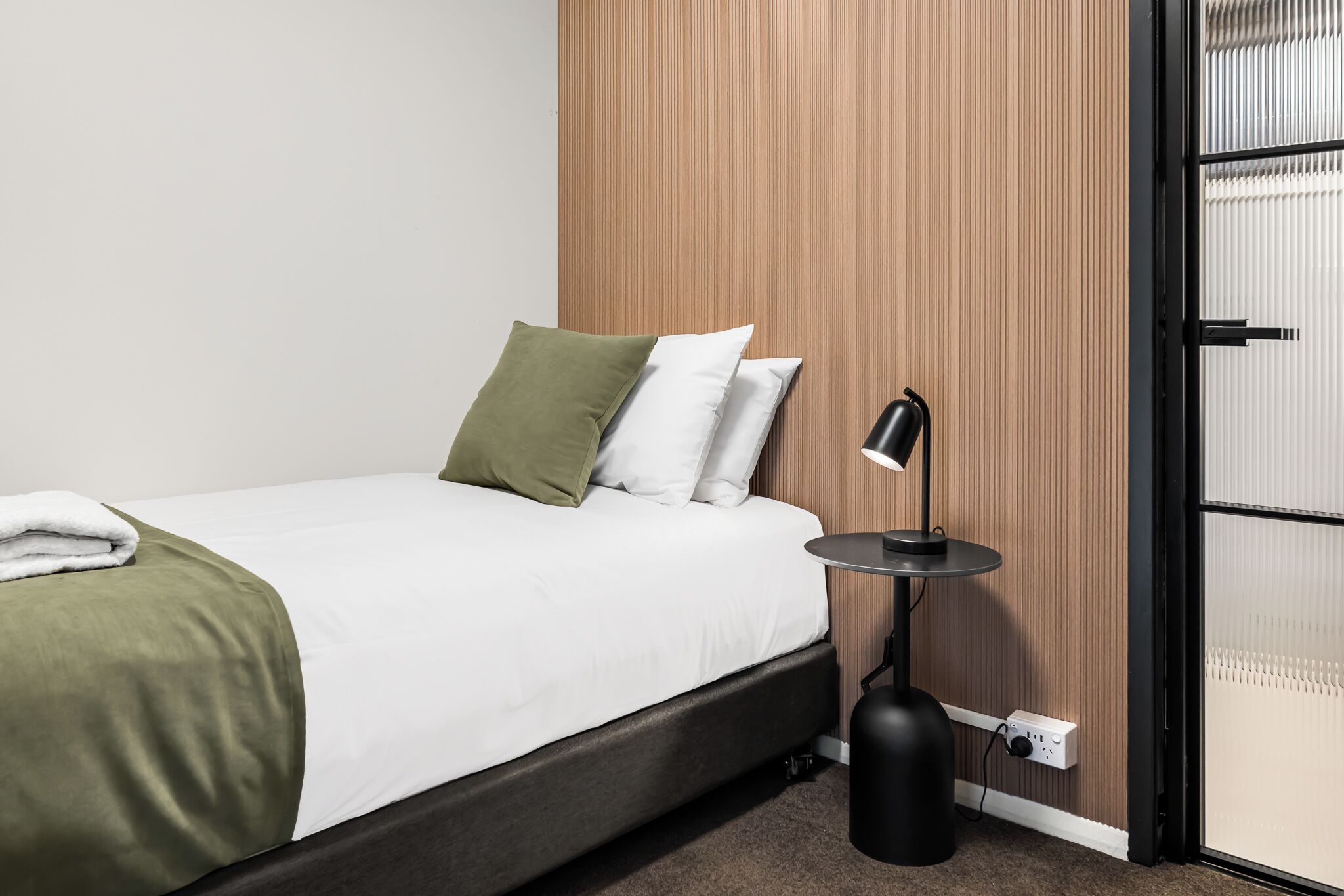 A neatly made bed with a green and white bedspread, a black bedside table with a lamp, and a wooden panel wall.