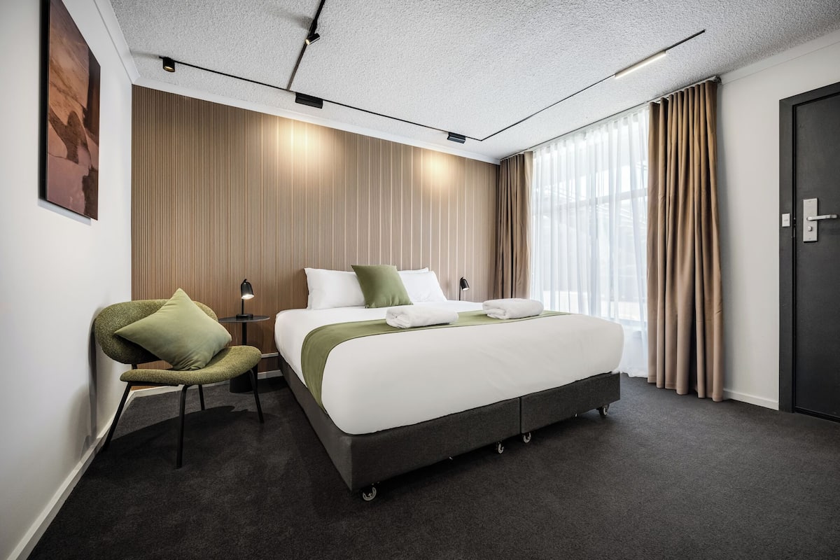 A modern hotel room with a large bed, a green chair, a desk with a lamp, and a dark door.