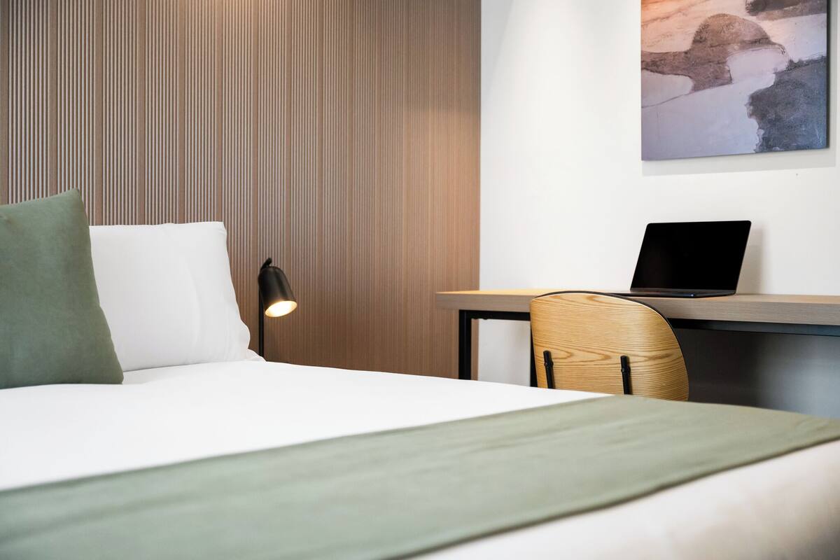 A hotel room with a bed, a desk with a laptop, a chair, and a wall-mounted painting.