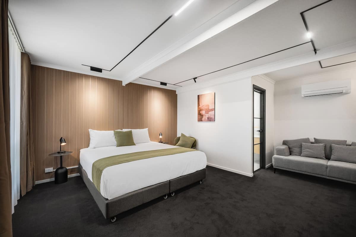 A modern hotel room with a large bed, a grey sofa, and a wooden accent wall.