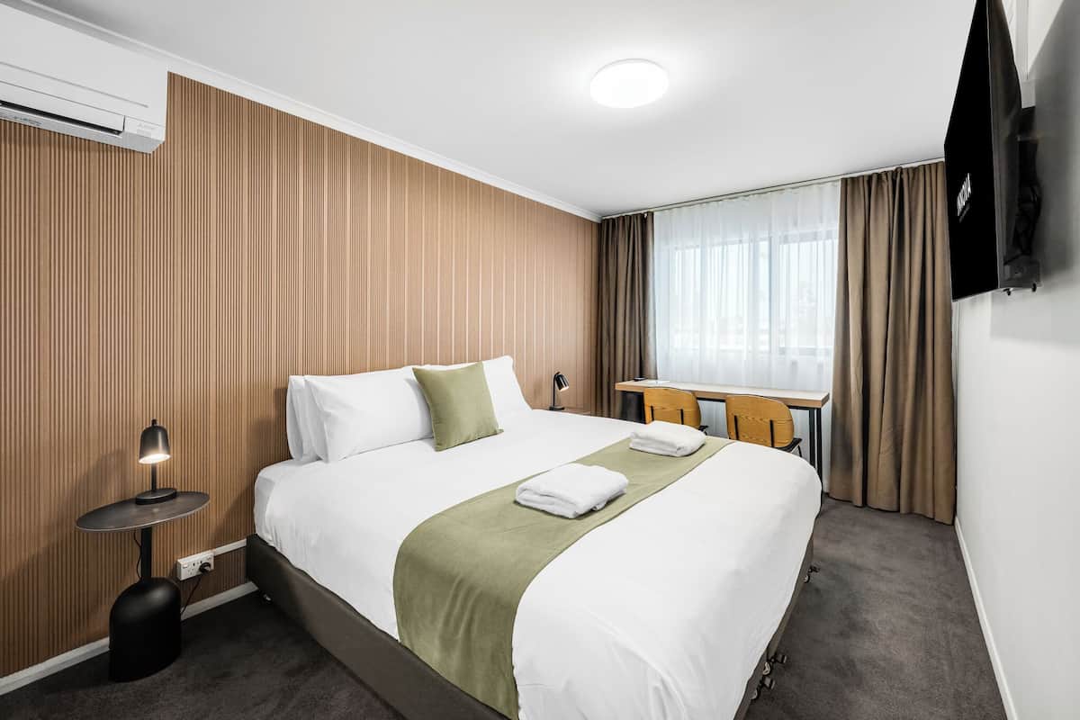 A hotel room with a bed, a desk, a chair, an air conditioner, and a television.
