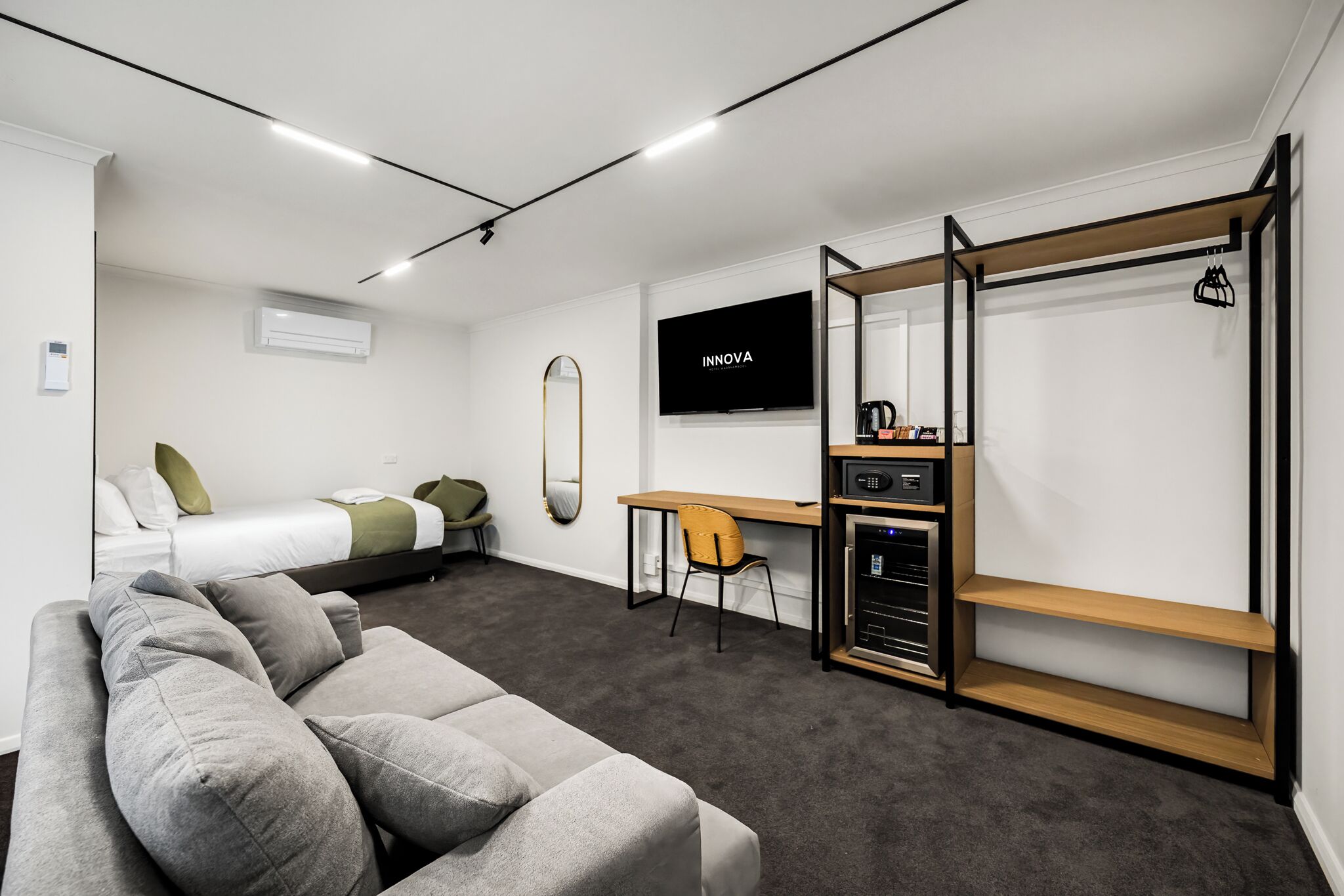 Habitación de hotel moderna y funcional, con un sofá gris, una cama con almohadas verdes, un escritorio de madera, un televisor y un estante fijado a la pared.