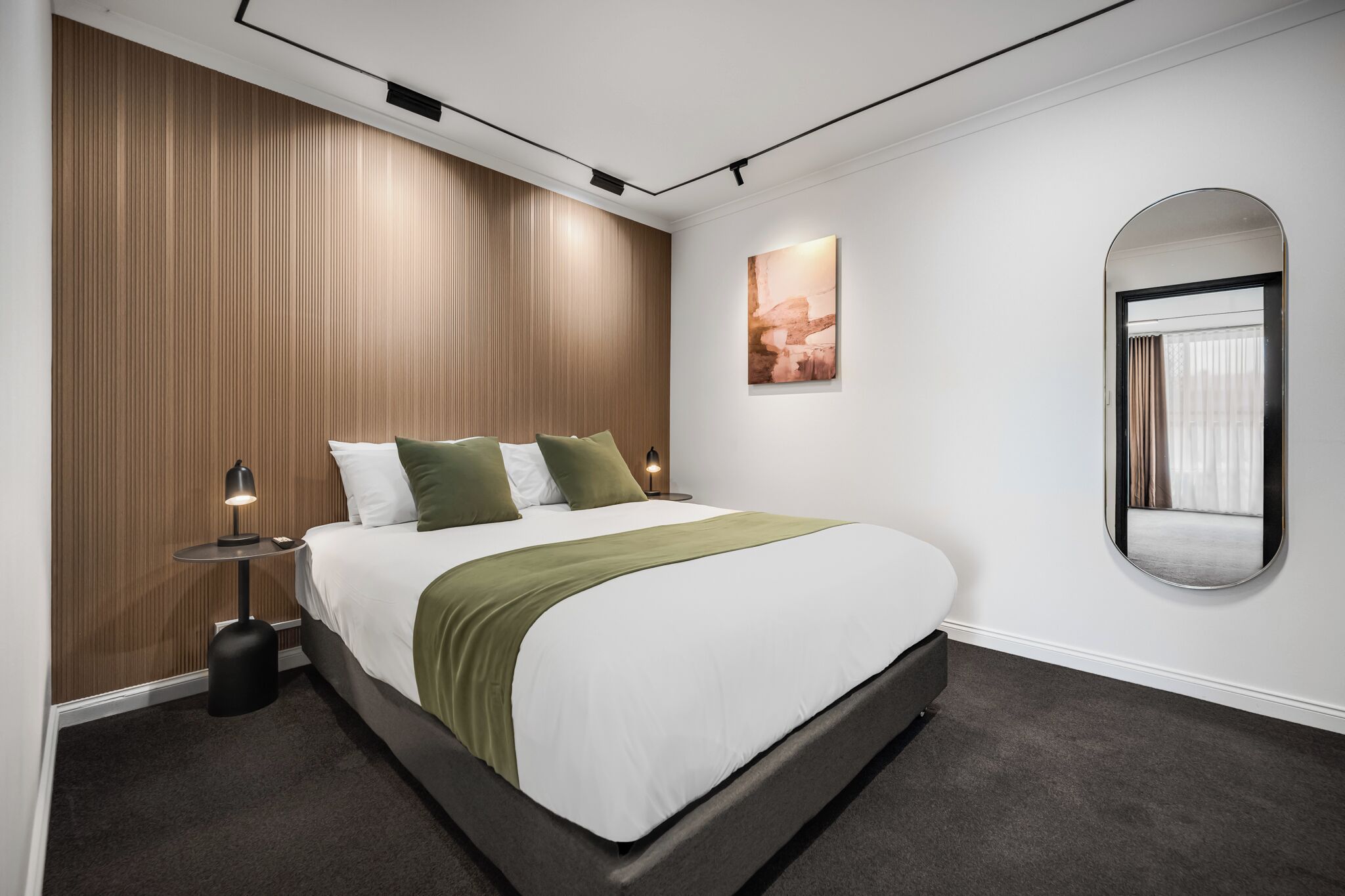 A modern hotel room with a large bed, a wooden accent wall, a small bedside table with a lamp, and a large mirror.