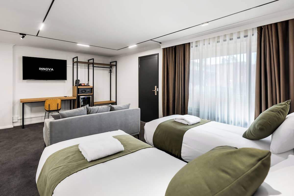 A hotel room with two beds, a desk, a TV, and a large window with curtains.