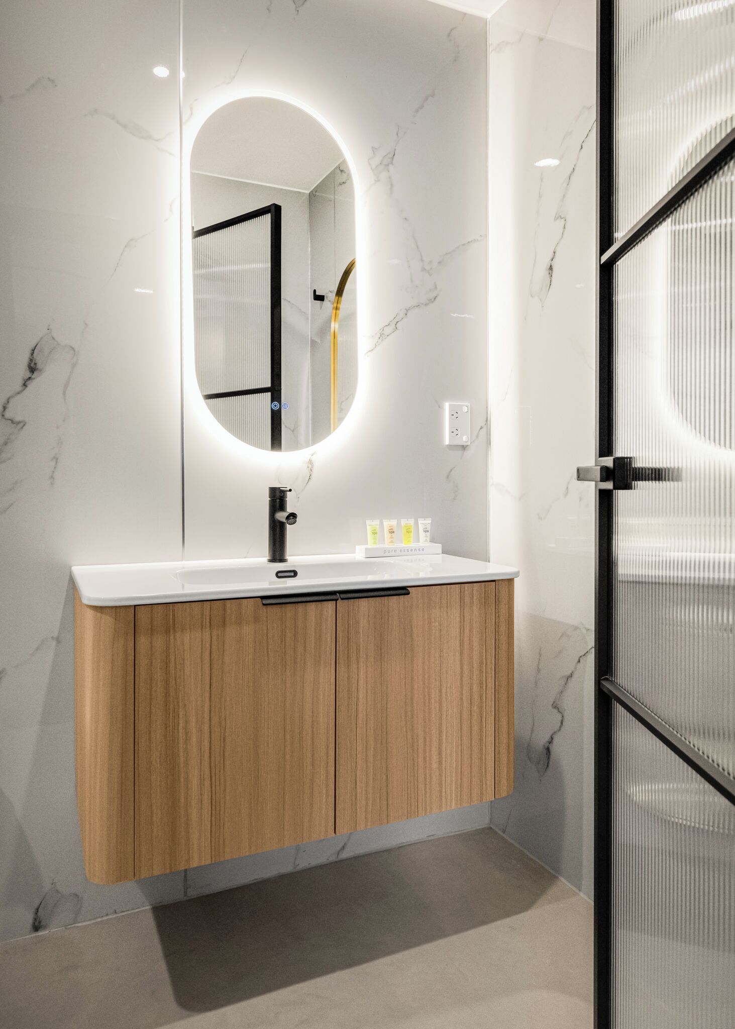 A modern bathroom with a marble wall, a wooden vanity, a sink with a black faucet, and a mirror with a circular light.