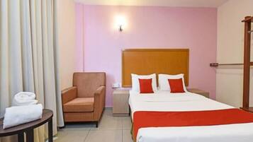 Quarto de hotel com cama, cadeira, mesa de cabeceira, luminária e janela com cortinas.