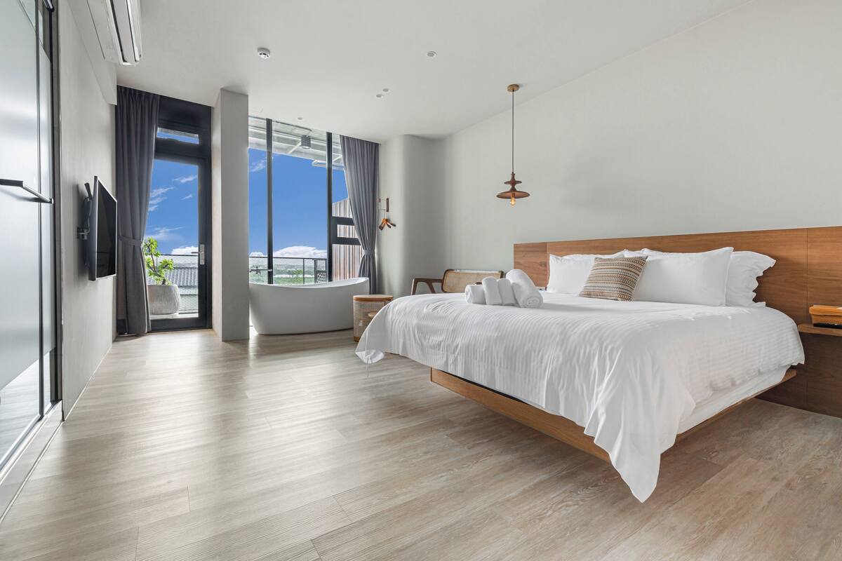 A modern hotel bedroom with a large bed, a view of the city, a bathtub, and a large window.