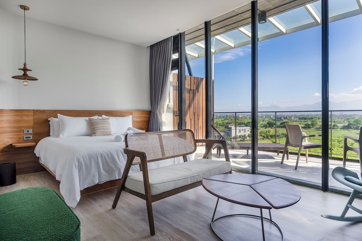 A modern hotel room with a large bed, a sofa, a small table, and a balcony with a view of the countryside.