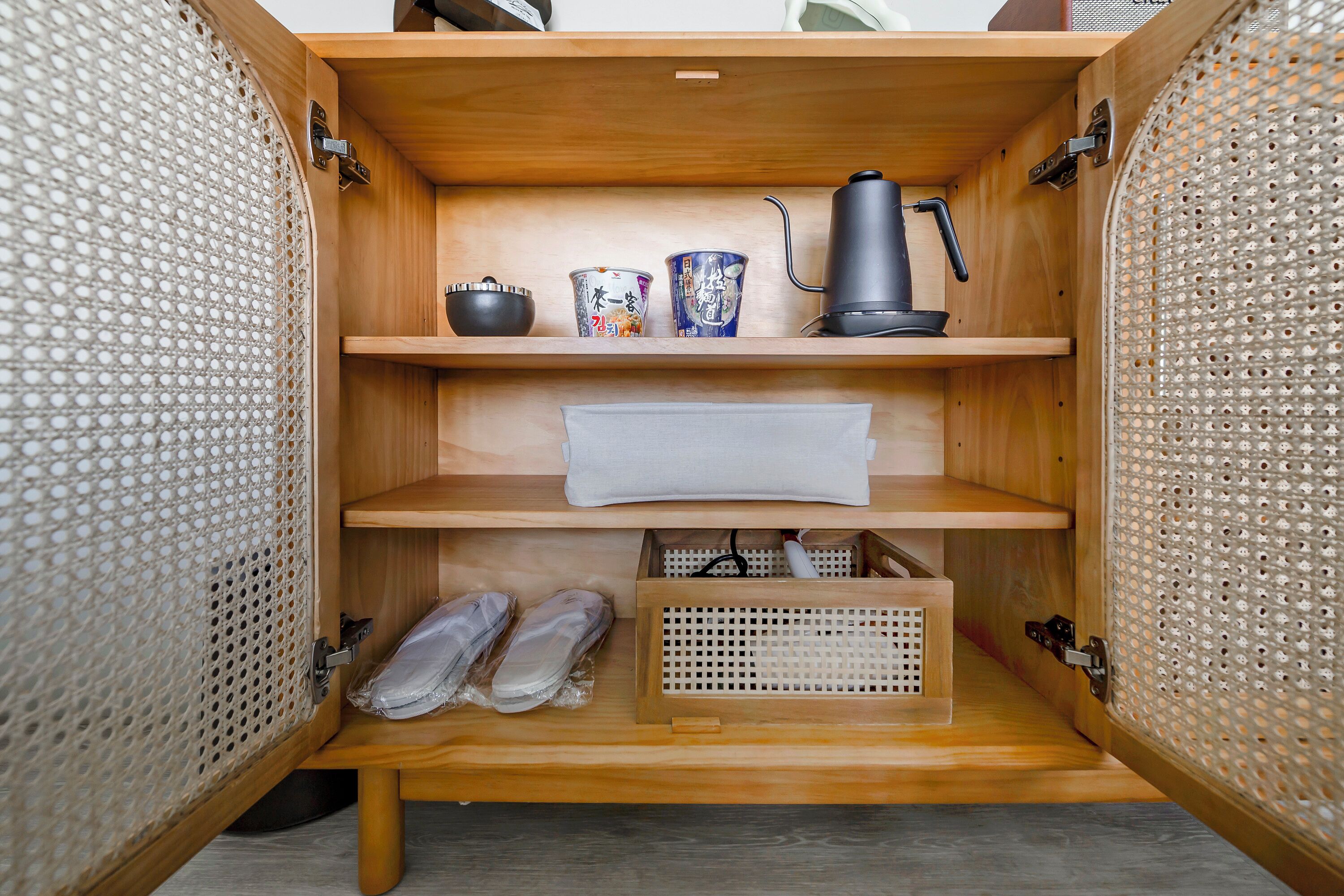 A wooden cabinet with shelves containing a kettle, cups, and a white towel.