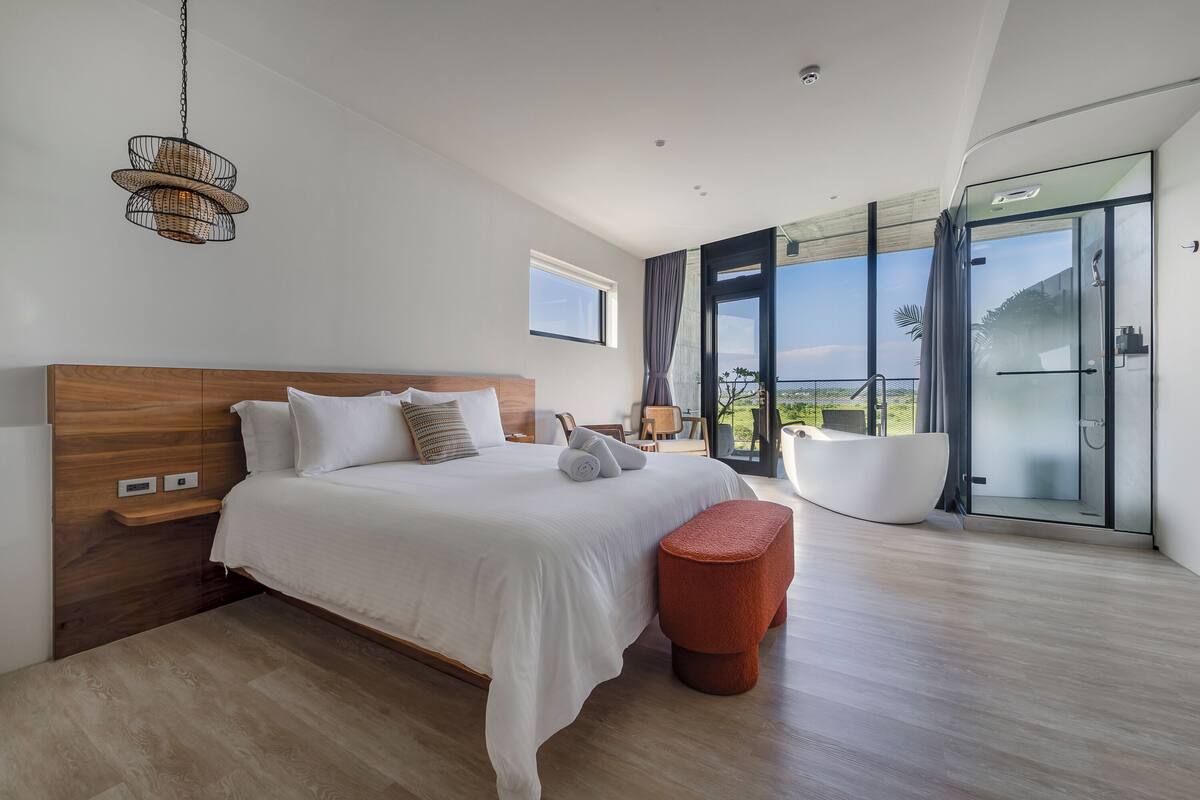 A modern bedroom with a large bed, a wooden headboard, a glass shower, and a view of the outdoors.