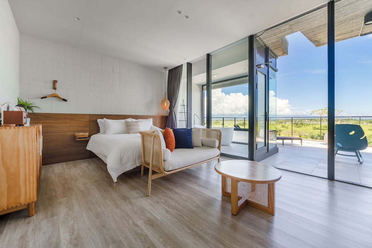 A modern bedroom with a bed, a wooden dresser, a round table, and a balcony with a view.