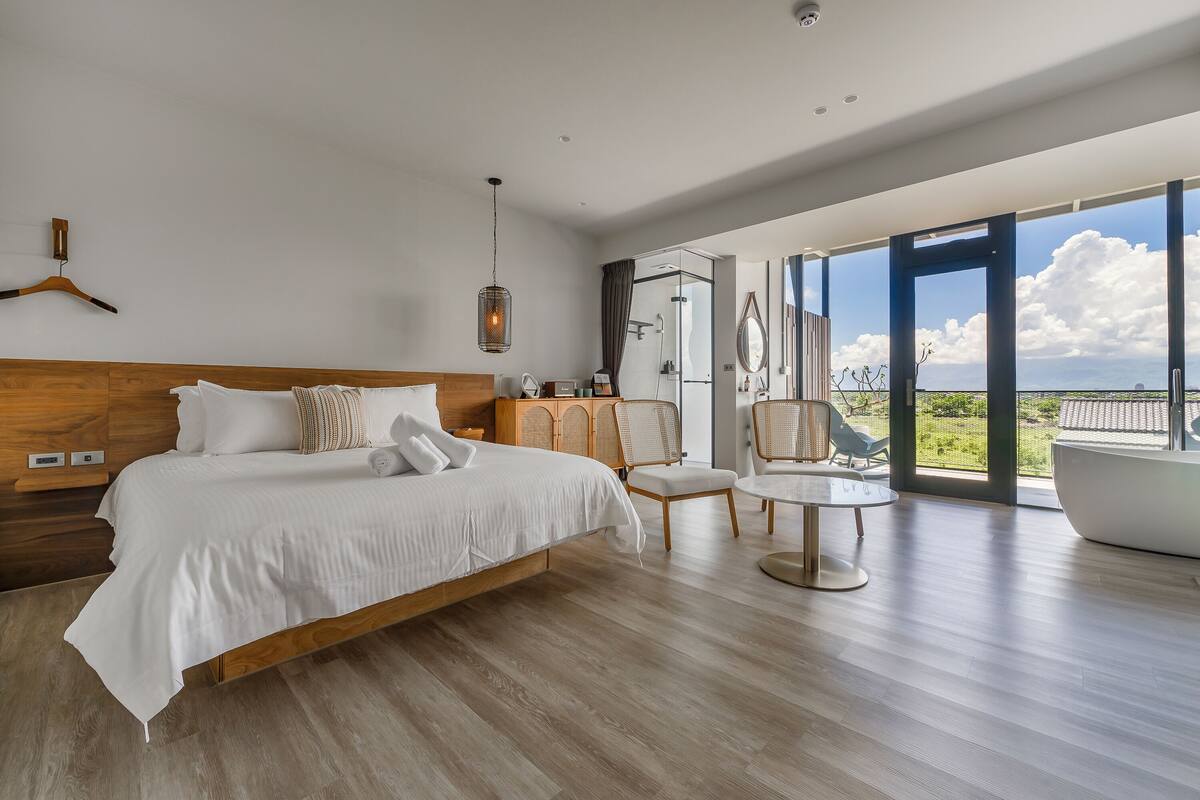 A modern bedroom with a large bed, a sitting area with chairs, a round table, and a bathtub.