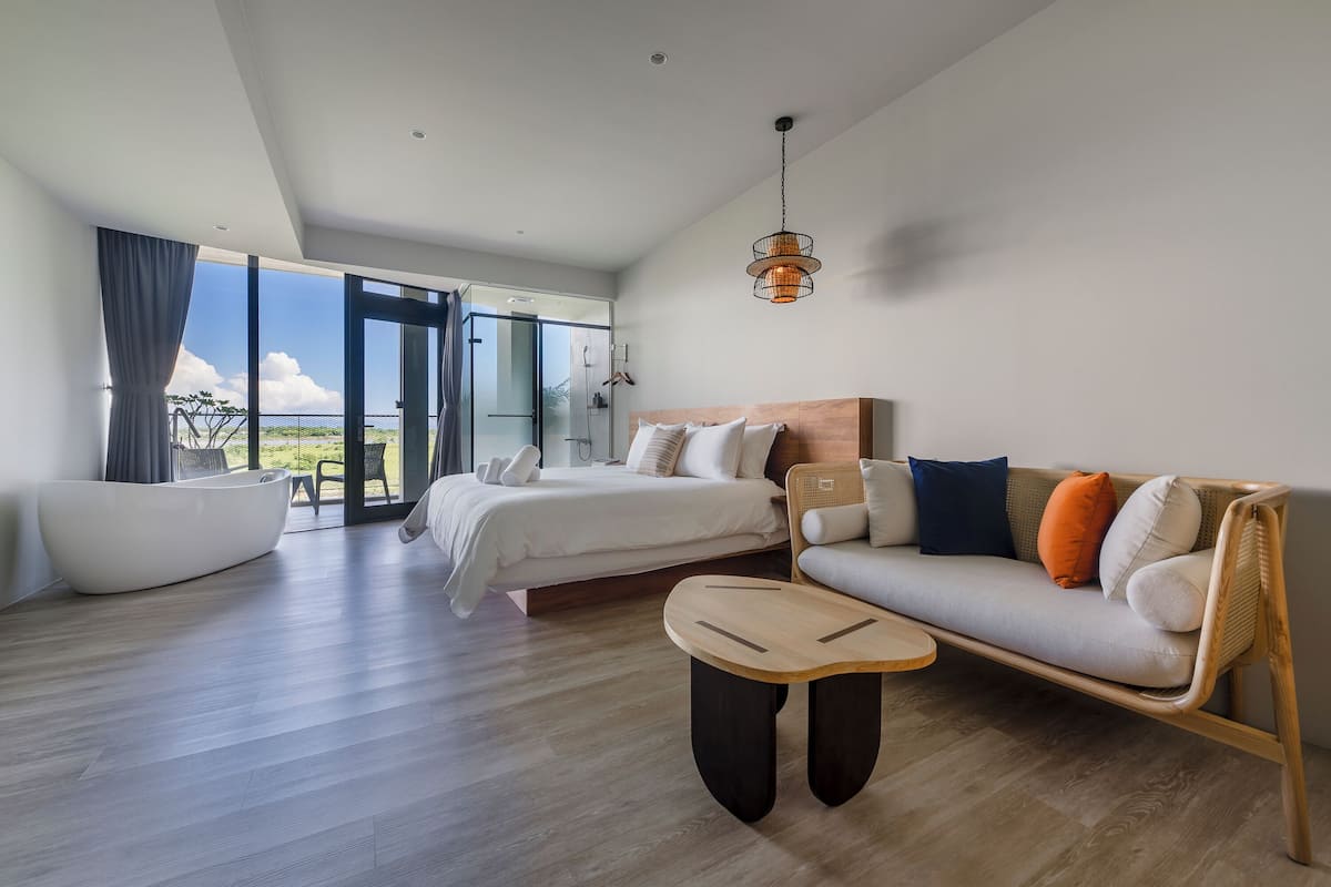 A modern hotel room with a large bed, a sofa, a wooden coffee table, and a view of the outdoors.