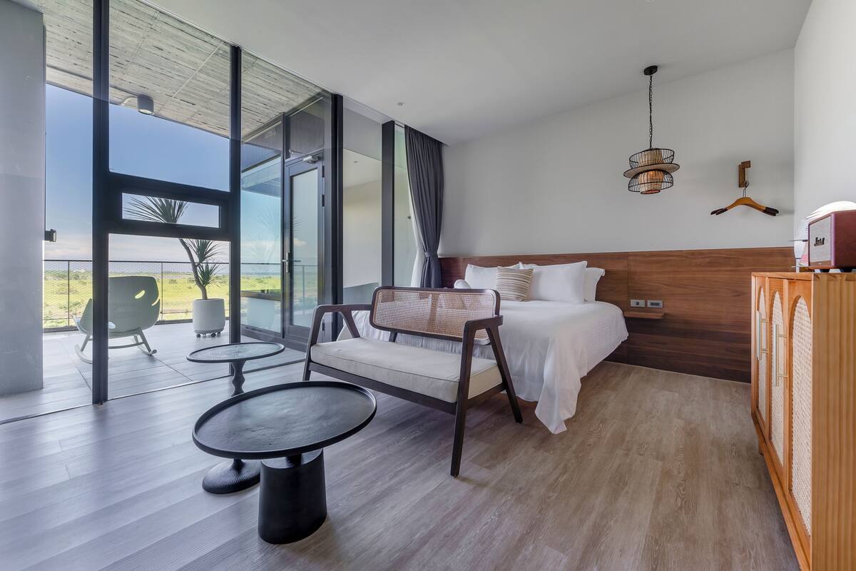 A modern bedroom with a large sliding glass door leading to a balcony, a bed with white linens, a wicker chair, and a small table.