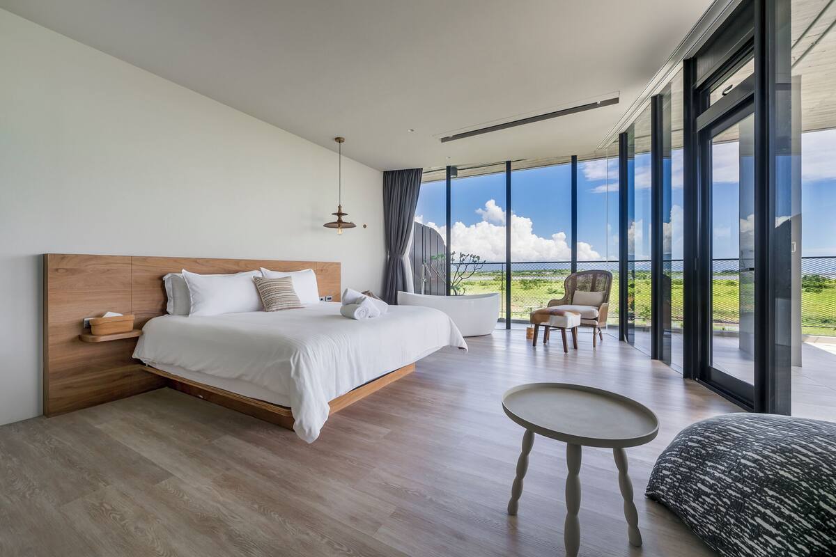 A modern bedroom with a large bed, a small table, and a chair. It has a view of the outdoors through large windows.