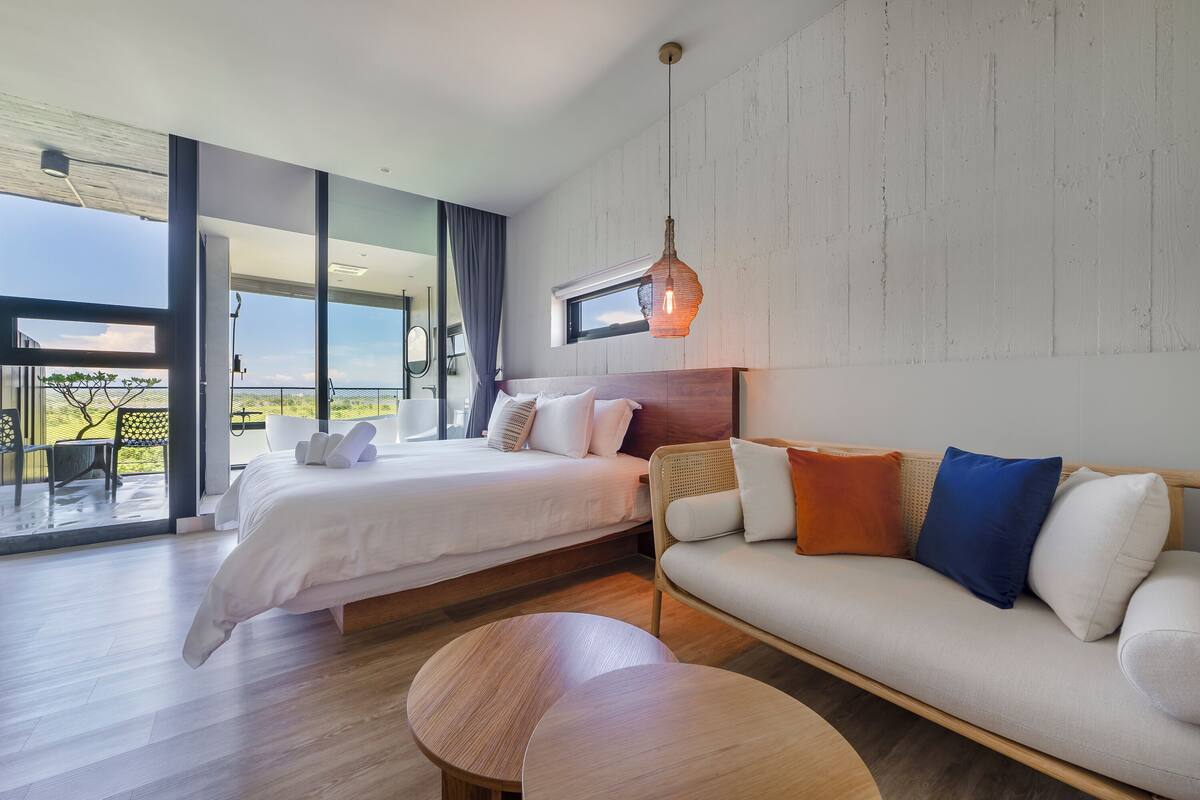 A modern bedroom with a large bed, a sofa, and a dining table. There is a balcony with a view of the landscape.