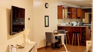A modern living room with a dining area, a kitchen with wooden cabinets, a flat-screen TV mounted on the wall, and a clock. - Sand Grains Residences 001 (Entebbe)