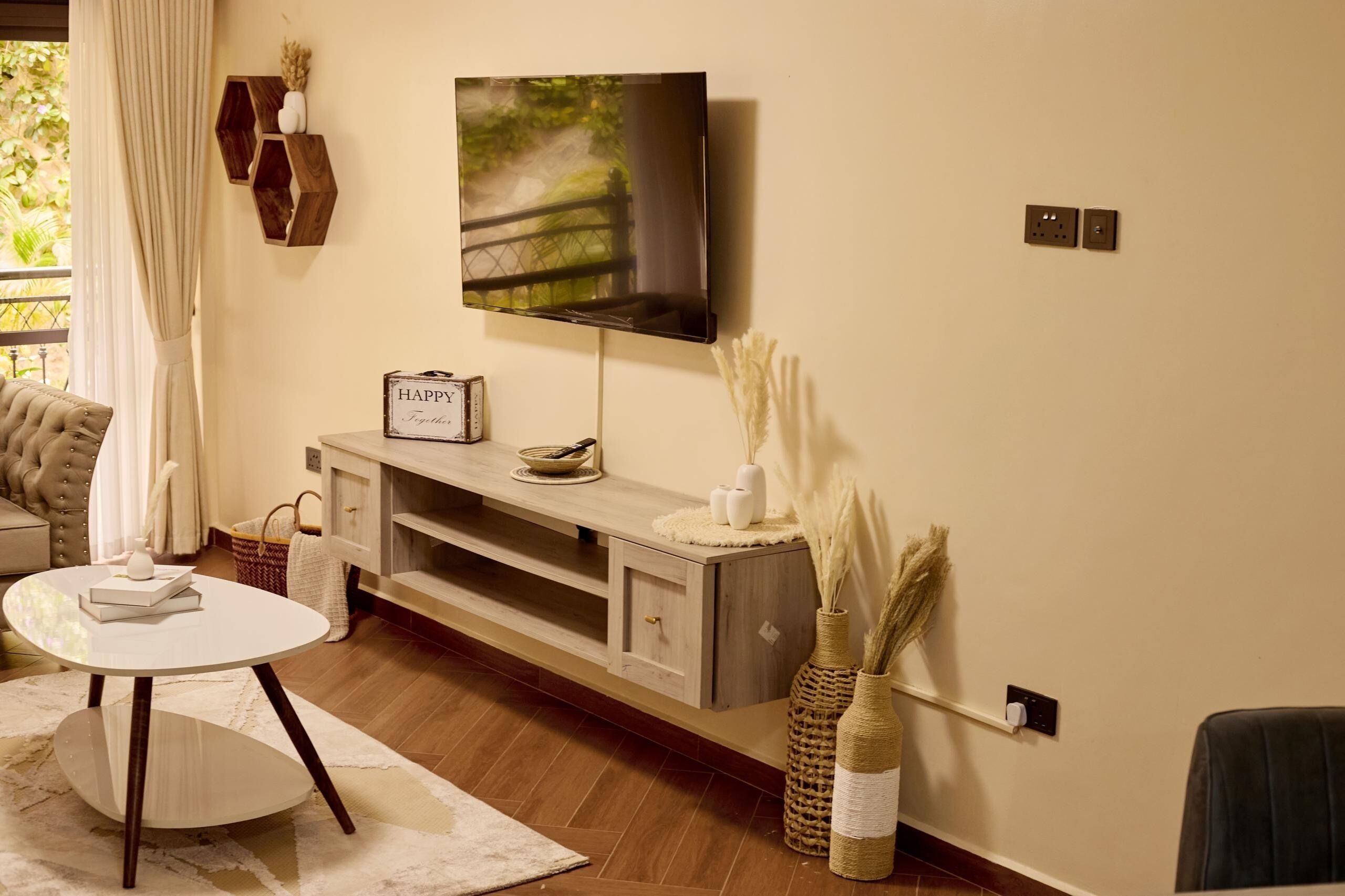 Un salon moderne avec un téléviseur à écran plat fixé au mur, une console en bois agrémentée d’objets décoratifs, une table ronde blanche et un canapé confortable.