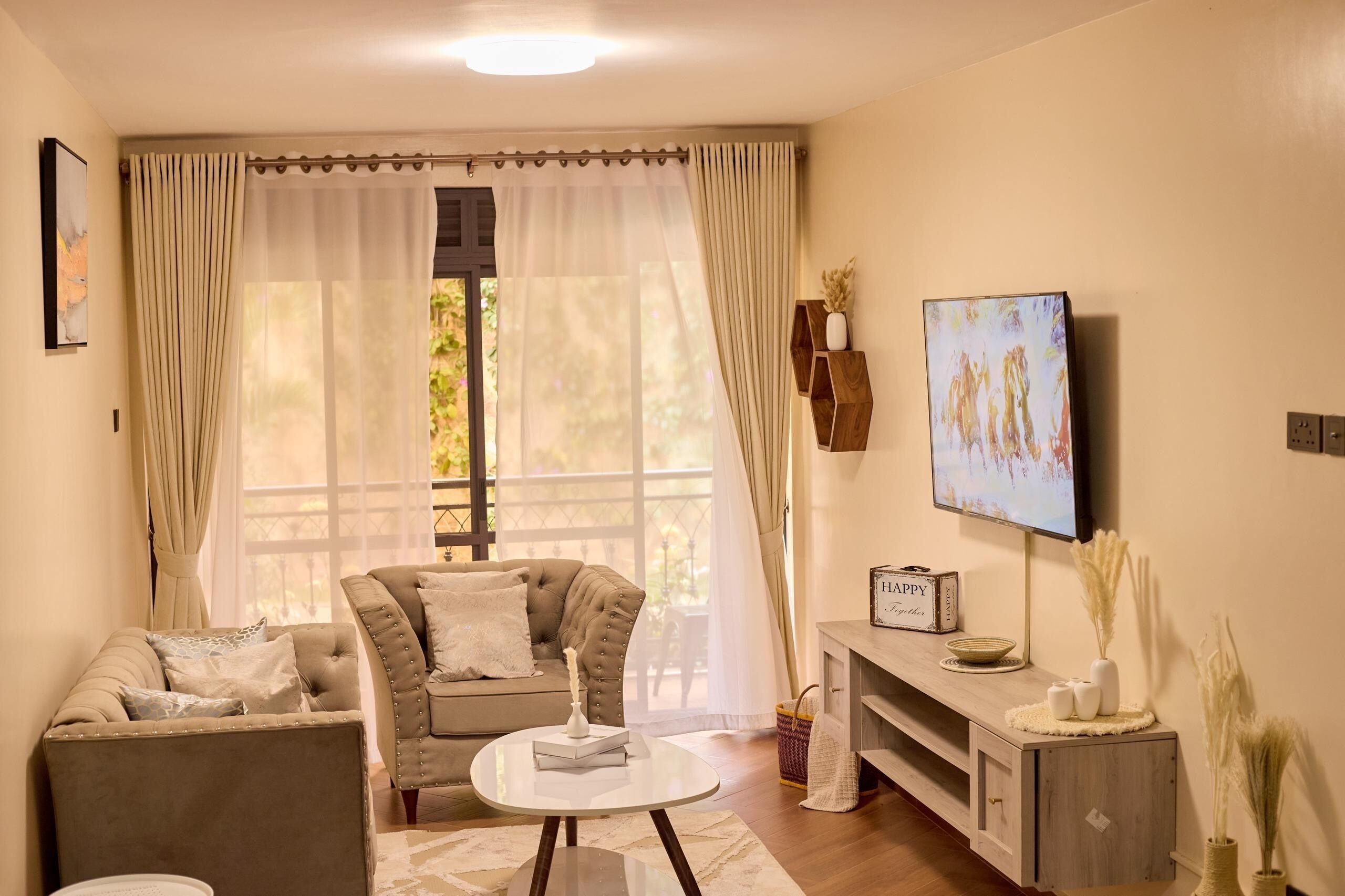 Een woonkamer met een bank, fauteuil en salontafel, een televisie aan de muur en een raam met gordijnen.