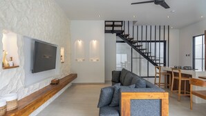 Een moderne woonkamer met een grijze bank, een houten console en een flatscreen televisie die aan een gestructureerde muur is bevestigd. Een trap met een zwarre leuning leidt naar een hoger gelegen verdieping.