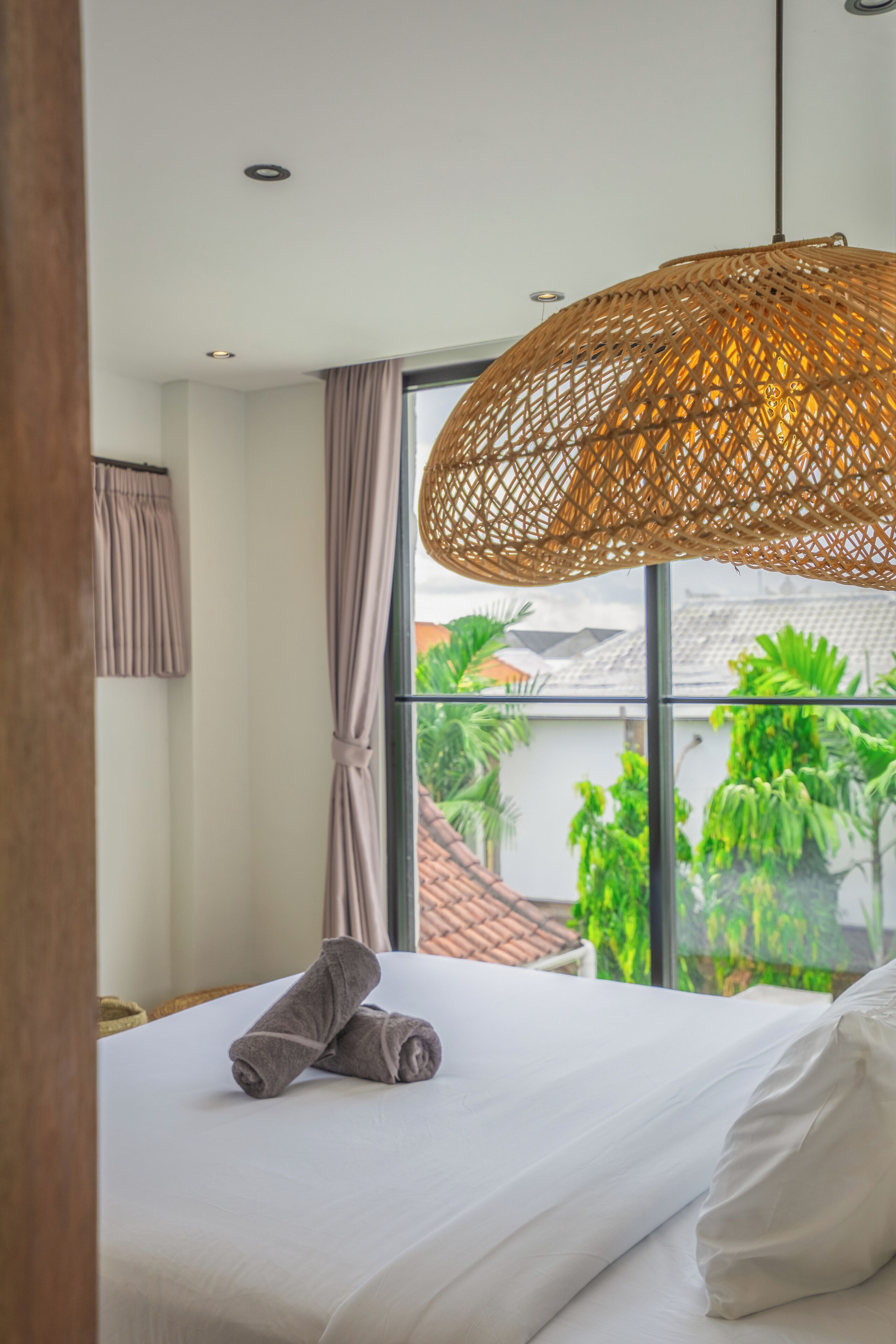 A hotel room with a large window, a bed with white linens, a rolled towel on the bed, and a woven light fixture.