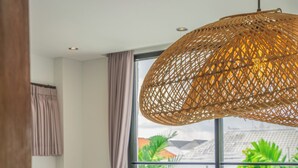 Een hotelkamer met een groot raam, een bed met witte lakens, een opgerolde handdoek op het bed, en een geweven lamp.