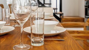 Een eettafel gedekt met transparant glaswerk en borden, een houten tafel en een trap op de achtergrond.