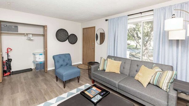 A living room with a grey sofa, blue armchair, and a coffee table. There is a window with curtains, a door, and wall decorations.