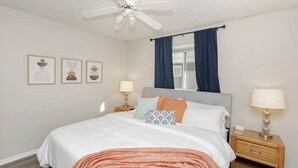 A bedroom with a bed, a ceiling fan, a window with curtains, bedside lamps, and wall art.