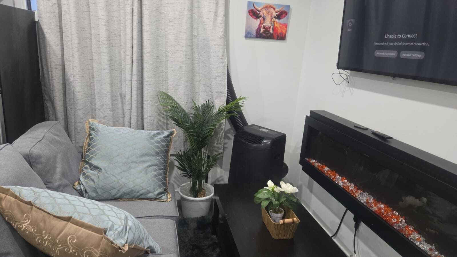 A living room with a grey sofa, a flat-screen TV mounted on the wall, a small table with a potted plant, and a decorative fish tank.