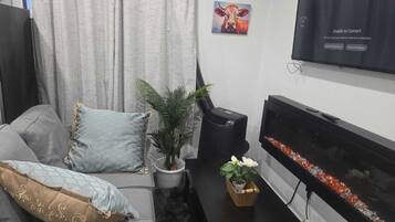A living room with a grey sofa, a flat-screen TV mounted on the wall, a small table with a potted plant, and a decorative fish tank.