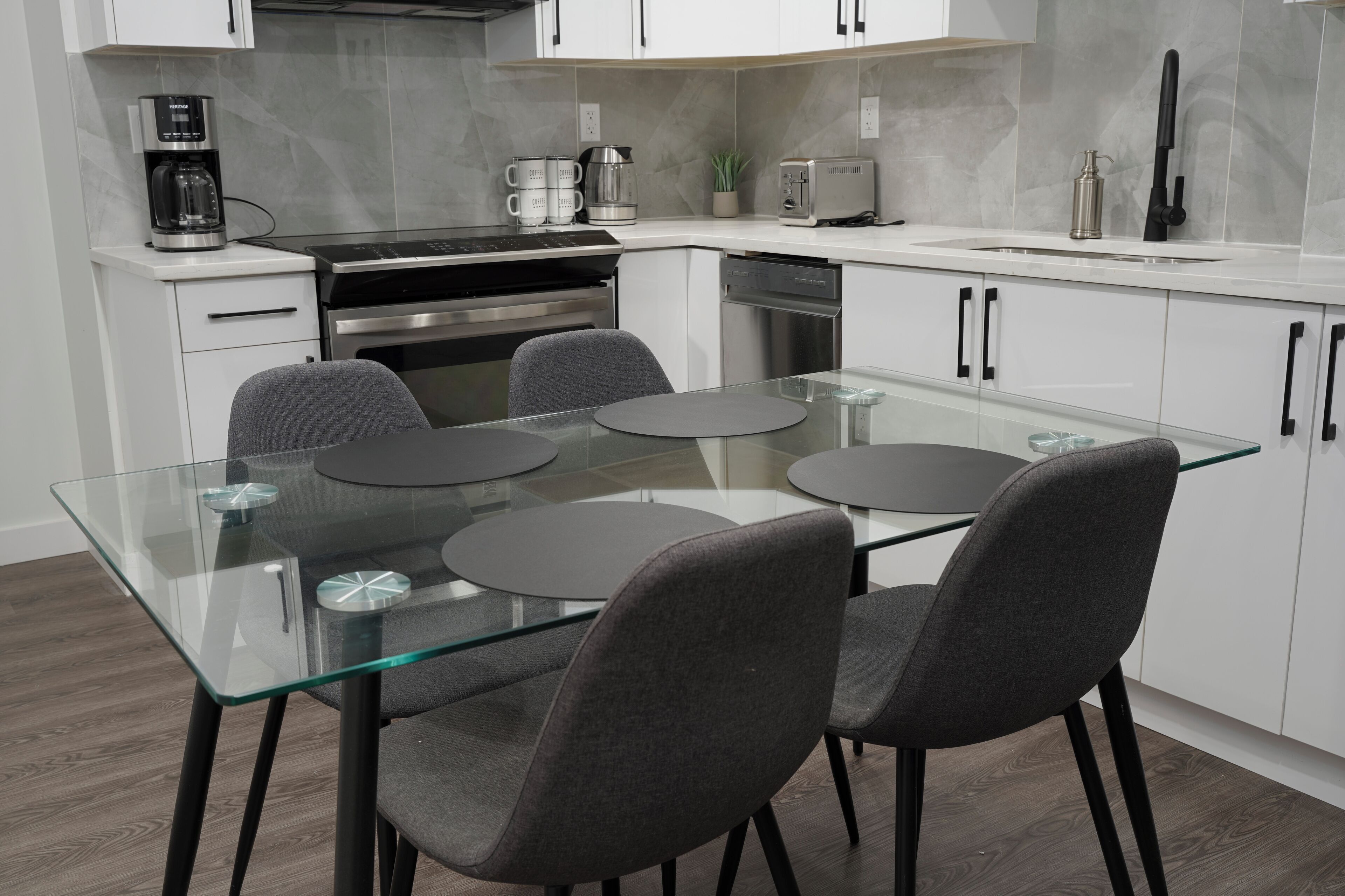 A modern kitchen with a glass dining table, four grey chairs, stainless steel appliances, and white cabinetry.