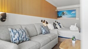 Una sala moderna con un sofá gris, cojines estampados, un panel de pared de madera, un cuadro de una playa y una pequeña mesa con una planta.