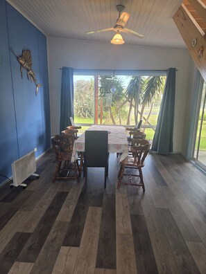 Un coin repas avec une table et des chaises, un ventilateur de plafond et une vue sur les palmiers à travers de grandes fenêtres.