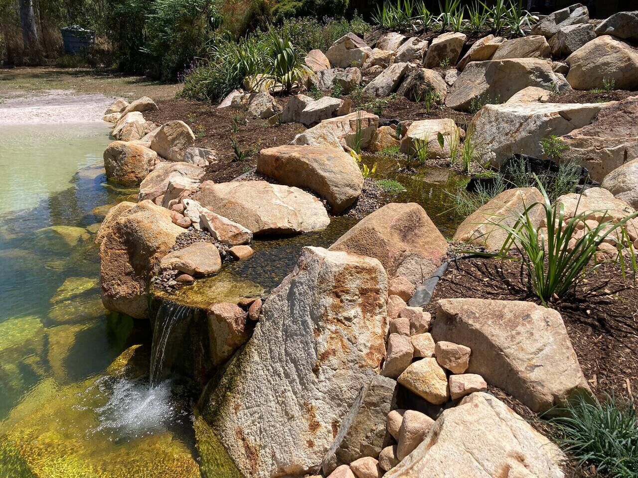 A natural rock formation with a small waterfall, surrounded by various plants and rocks.