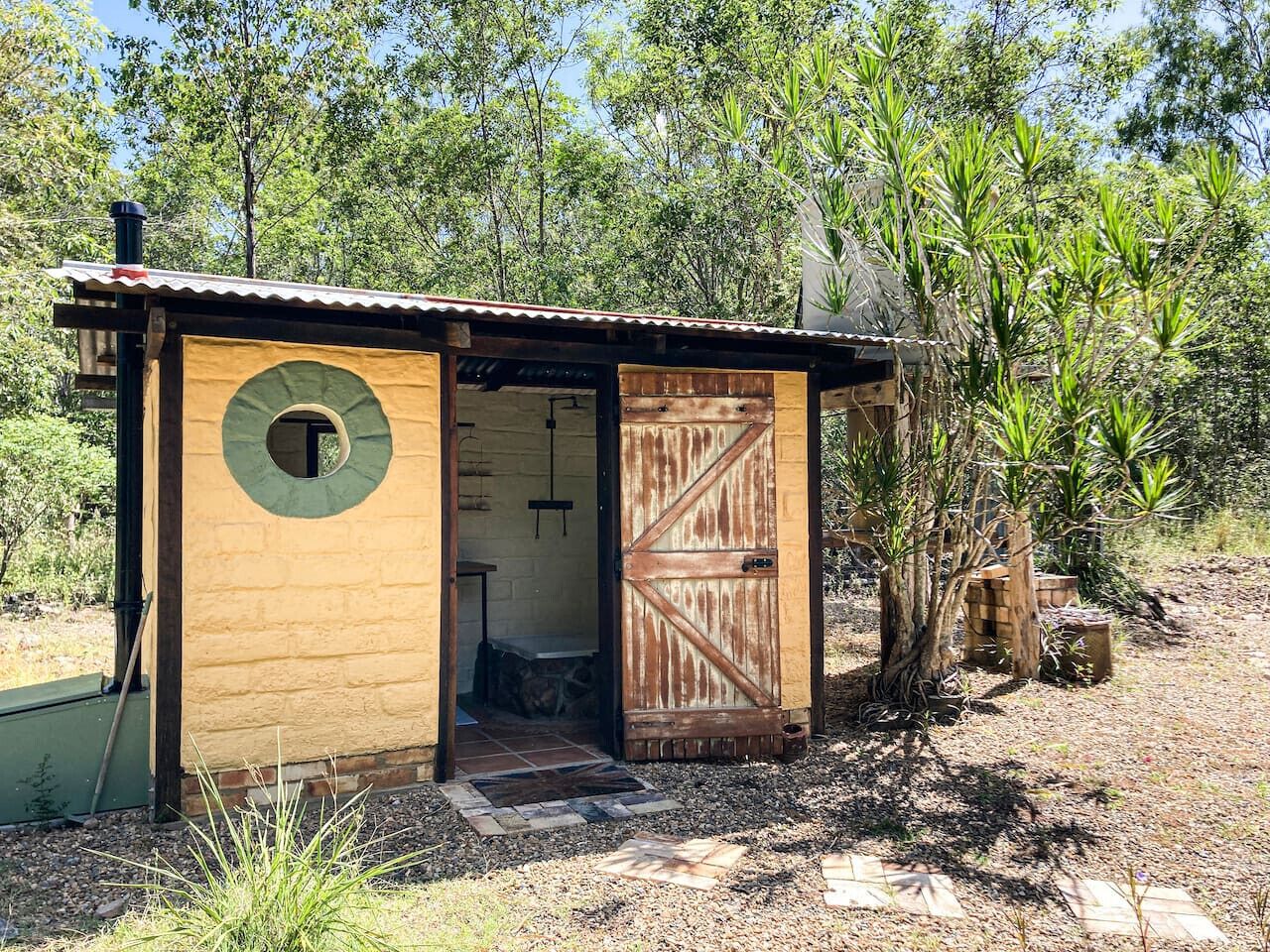Une cabane rustique en bois, dotée d’une fenêtre ronde et d’une porte en bois, entourée d’une végétation naturelle.