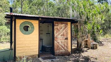Une cabane rustique en bois, dotée d’une fenêtre ronde et d’une porte en bois, entourée d’une végétation naturelle.