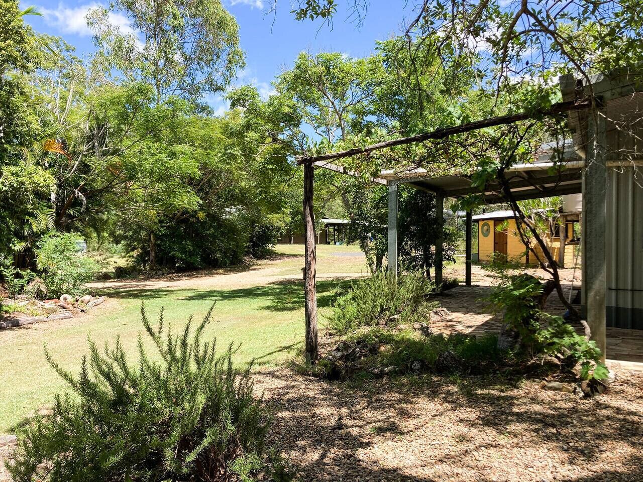 Um jardim com pérgola de madeira, gramado e um edifício com fachada amarela.