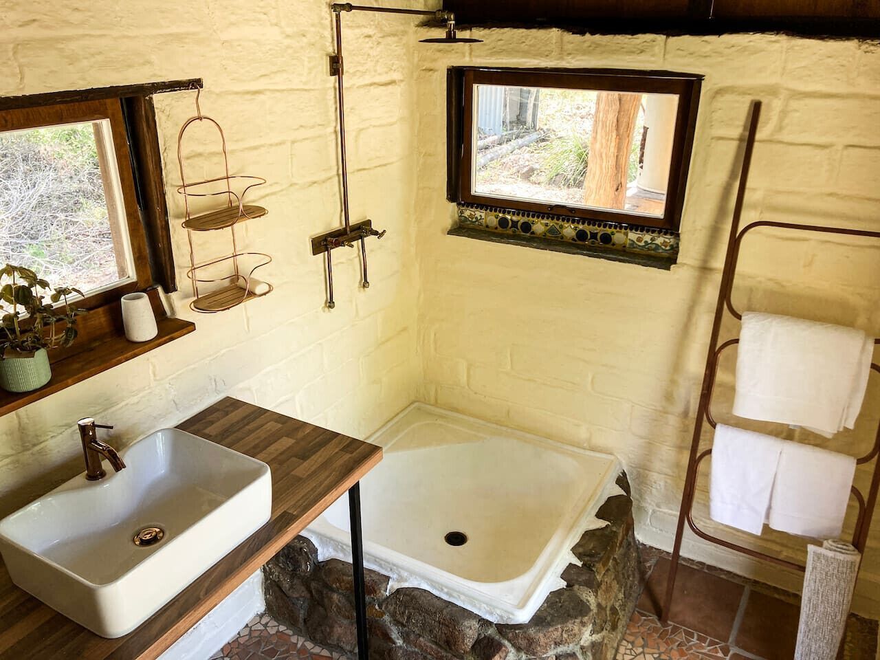 Un baño con tocador de madera, lavamanos blanco, ducha y perchero para toallas.